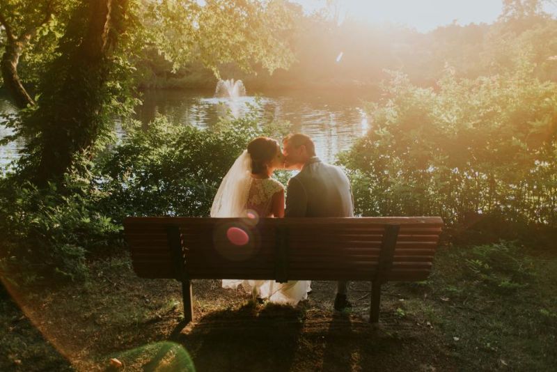 A couple kissing on a park bench in warm sunlight at their wedding.